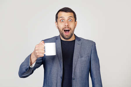 Young businessman man with a beard in a jacket, holding a cup of coffee, scared in shock with a surprised face, scared and excited with an expression of fear. Portrait of a man on a gray background.の写真素材