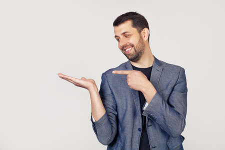 Young businessman man with a beard in a jacket looks at the palm, showing with his hand and pointing his finger. Portrait of a man on a gray background.の写真素材