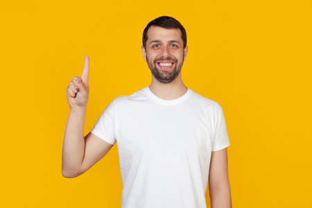 Modern young man with a beard in a white tank top shows number one with fingers on hand, smiling confidently and happily, looking at the camera. The man shows one finger. Number 1, yellow backgroundの写真素材