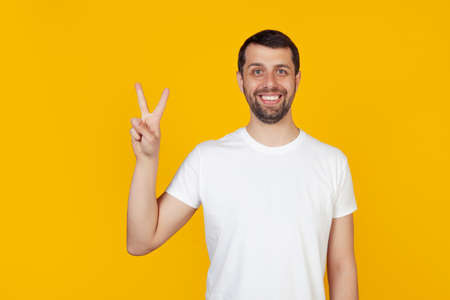 Modern young man with a beard in a white tank top shows number two with fingers on hand, smiling confidently and happily, looking into the camera The man shows two fingers. Number 2 yellow background.の写真素材
