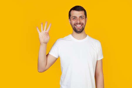 Modern young man with a beard in a white tank top shows number four with fingers on hand, smiling confidently and happily, looking at the camera The man shows four fingers. Number 4.yellow backgroundの写真素材