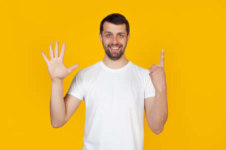 Modern young man with a beard in a white tank top shows number six with fingers on hand, smiling confidently and happily, looking into the camera The man shows six fingers. Number 6.yellow backgroundの写真素材
