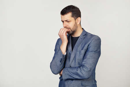 Young businessman with a smile, a man with a beard in a jacket, looks tense and nervous with hands on his lips, biting his nails. Anxiety problem. Portrait of a man on a gray background.の写真素材