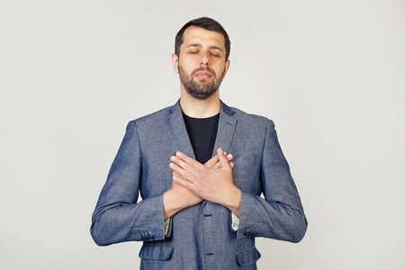 Young businessman with a smiling man with a beard in a jacket, with hands on his chest with closed eyes and a grateful gesture on his face. Health concept. Portrait of a man on a gray background.の写真素材