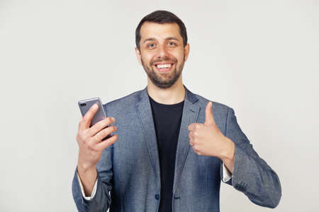 Young businessman with a smile, a man with a beard in a jacket, with a smartphone in his hand, happy with a big smile, makes an OK sign, thumb up with fingers, a great sign.の写真素材