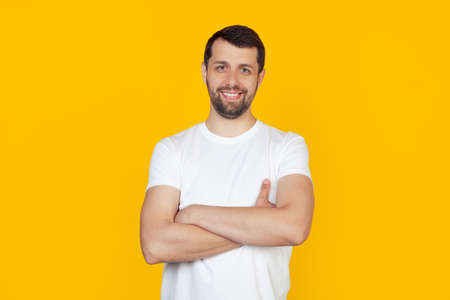 Young man with beard in white t-shirt happy face smiling with crossed arms looking at the camera. Positive man stands on isolated yellow background.の写真素材