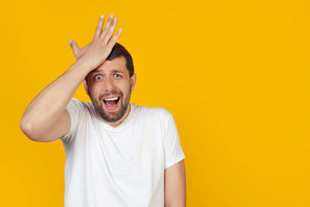 Young man with a beard in a white t-shirt, surprised, put his hand on his head for a mistake,...