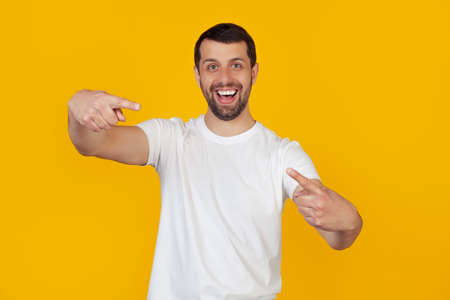 Young man with a beard in a white t-shirt pointing with his fingers at the camera with a happy and funny face. Good energy and vibes. Stands on isolated yellow background.の写真素材