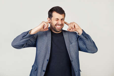 Young businessman man with a beard in a jacket covering his ears with his fingers with an annoyed...