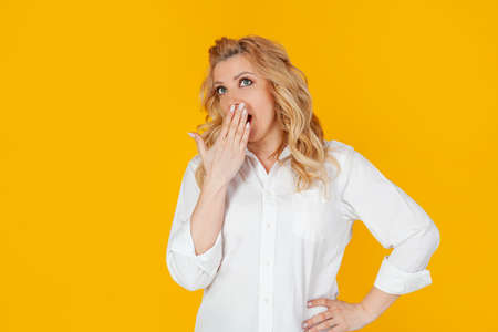 Portrait of a blandin woman in a white shirt, middle-aged European girl yawning and covering her open mouth, with a boring face, wakes up early, needs coffee, stands against a yellow background.の写真素材
