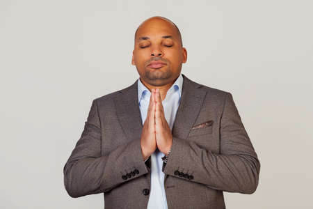 Handsome young concentrated african american businessman male in jacket holding hands together in front of face, eyes closed praying and hoping today is a good day, on gray backgroundの写真素材