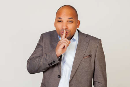 Man african american businessman in a jacket makes a gesture with his hand holding a finger on his lips, asks for silence and silence.の写真素材