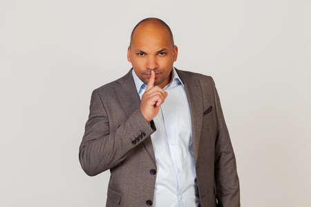 Portrait of a young African American guy businessman holding a finger on his lips, shows to close his mouth and stop making noise at the wrong moment. Standing on a gray backgroundの写真素材