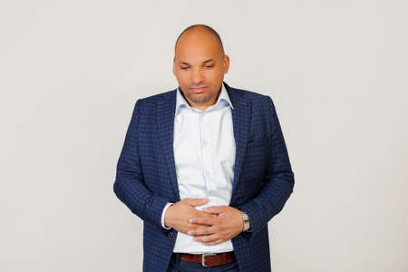 Portrait of a sick young African American businessman guy, with a hand on his stomach due to indigestion, feeling sick. Pain concept. Standing on a gray backgroundの写真素材