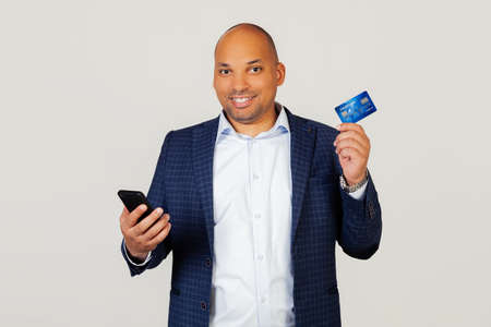 Portrait of happy young African American guy businessman using credit card to pay online using smartphone. Standing on a gray backgroundの写真素材