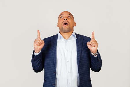 Portrait of astonished young African American businessman guy, amazed and surprised, looking up and pointing with fingers and raised hands. Standing on a gray backgroundの写真素材
