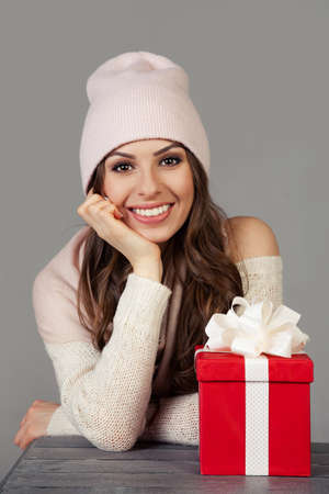 Smiling and happy, beautiful young woman in winter clothes with a red Christmas gift box on a gray background. The girl enjoys Christmas shopping, meets the New Year. Shopping sale concept.の写真素材