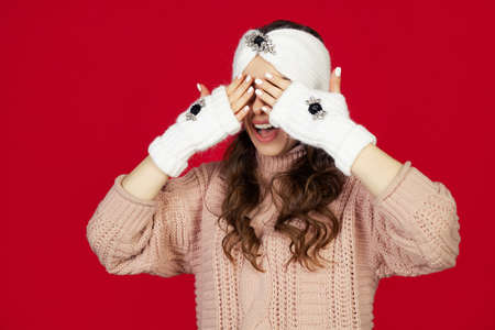 Smiling and happy, beautiful young woman in winter clothes covering her eyes with her hands, waiting for a Christmas present. New Years surprise. Standing on a red background.の写真素材