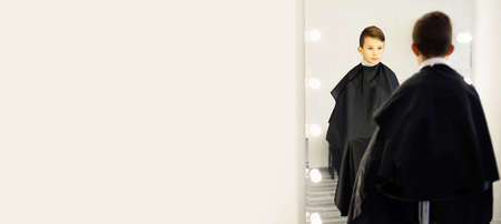 A 10-year-old boy sits in a barbershop chair in front of a mirror. A young child in a beauty salon sits with a black cape, looks in the mirror .の写真素材