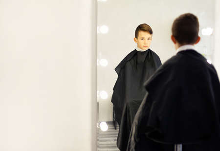 A 10-year-old boy sits in a barbershop chair in front of a mirror. A young child in a beauty salon sits with a black cape, looks in the mirror .の写真素材