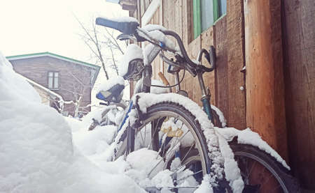 Bicycles covered with snow stand against a wooden wall near the house. Old abandoned bicycles in the backyard of a house under the snow.の写真素材