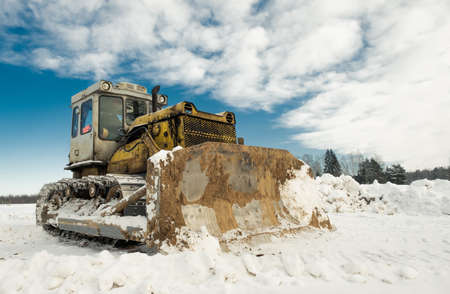 Yellow crawler tractor bulldozer with a bucket works in winter clearing the road from snow. Frosty day with bright sun and beautiful blue sky in the background. Yellow bulldozer in winter.の写真素材
