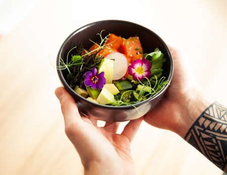 A man holds a bowl of Poke salad with salmon, avocado, cucumber, rice, Tamarind sauce, mango sauce,...