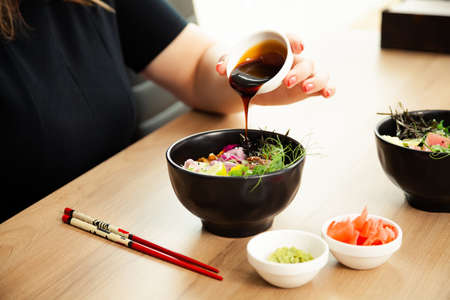 Girl adding teriyaki sauce to Poke salad. Dressing salad with sauce in a restaurant or cafeの写真素材
