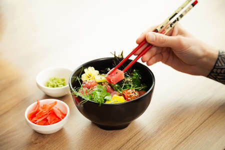 A man eats a Poke salad with tuna in a bowl with chopsticks. Place the tuna salad in a bowl. Young man guy in a restaurant eating poke salad. Asian seafood salad conceptの写真素材
