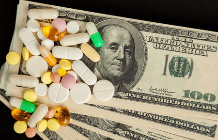 Covid-19 coronavirus pills are scattered across the hundred dollar bills. A handful of pills on...