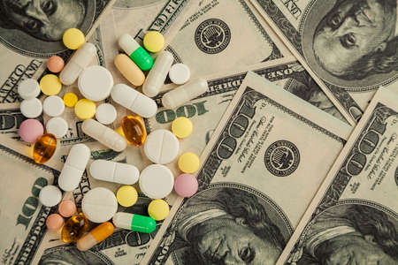 Covid-19 coronavirus pills are scattered across the hundred dollar bills. A handful of pills on...