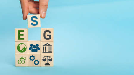 ESG concept of environmental, social and governance. Words ESG on a wood cubes. Hand holds wooden cube with abbreviation ESG standing with other icons on blue backgroundの写真素材