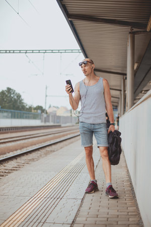 Young man in a t-shirt on the platform waiting for a train using mobile phone. Man by train station platform suburban with bag and mobile phone. Traveling by train.の写真素材