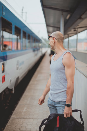 Young man in a t-shirt on the platform waiting for a train using mobile phone. Man by train station platform suburban with bag and mobile phone. Traveling by train.の写真素材