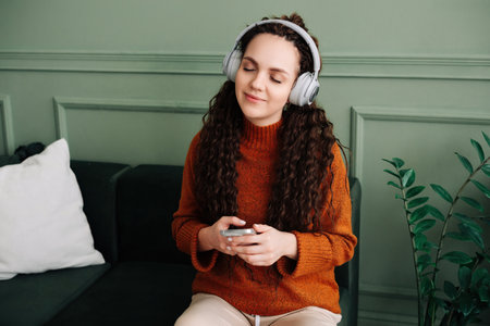 A calm, relaxed, carefree young woman listens to music, an audio podcast or audiobook on her cell phone while sitting at home, meditating and relaxing, feeling peace of mind with her eyes closed.の写真素材
