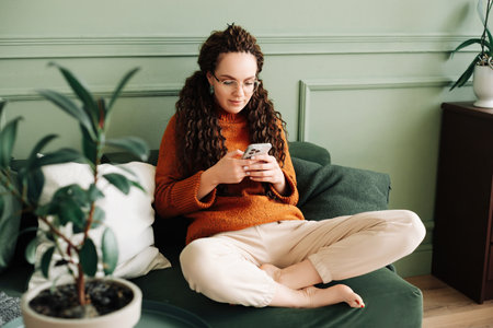 Relaxed young woman sitting on a sofa using a cell phone at home holding a smartphone, looking at cell phones shopping in mobile apps ordering e-commerce products, or checking social media.の写真素材