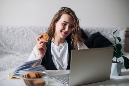 Young redhead woman watching series or podcast looking at screen with interest holding cookie in hand sitting at kitchen table with glass jar and laptop, smiling happily. Unconscious eating disorderの写真素材