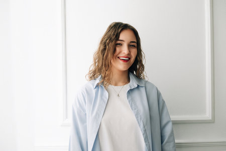 Confident young woman in casual blue tee smiling at camera by window, against white wall. Playful and vibrant indoor portrait.の写真素材