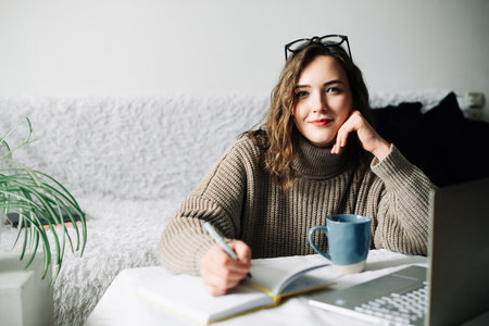 Stylish and focused female PR manager creating content plan for online store, working from home with laptop, notebook and coffee, looking at cameraの写真素材