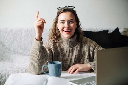 Eureka Excited female entrepreneur with successful business idea, sitting at kitchen table with laptop and showing index finger, ready to make a brilliant project and business planの写真素材