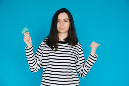 Woman Holding Credit Card, Pointing with Thumb in Empty Space - Female Expressing Confidence and Financial Freedom - Blue Background - Perfect for Banking, E-commerce, and Financial Services Conceptsの写真素材