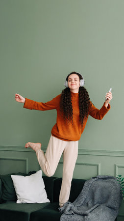A happy, pretty young woman wearing headphones dances to her favorite song at home. Cheerful funny girl listening to popular music, moving, jumping on the couch in the living room, experiencing joy.の写真素材