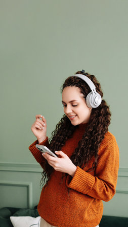 Happy pretty young woman wearing headphones dancing at home. Cheerful girl listening to music on mobile phone, singing songs feeling relaxed standing in modern living room interior.の写真素材