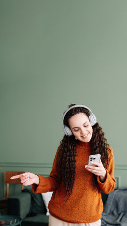 Happy pretty young woman wearing headphones using a smartphone dancing at home. Cheerful girl listening music on mobile phone, singing song feeling relaxed standing in modern living room interior.の写真素材