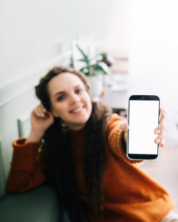 Happy young woman holding mobile phone in hand showing cellphone display, presenting big white blank cellular display template mock up, smartphone mockup screen for digital application promo ads.の写真素材