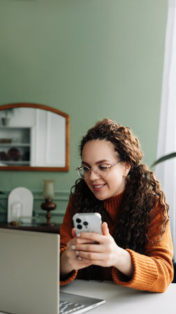 Smiling Young woman using smartphone for remote work and e-learning at home. Modern Technology: Young Woman Using Smartphone for Work and Learning at Home. Pretty woman holding mobile phoneの写真素材