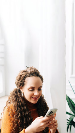 Young woman browsing online on cellphone while sitting comfortably. Happy woman shopping on smartphone while relaxing on couch. Smiling lady using smartphone for social media and e-commerceの写真素材