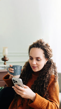 Young woman enjoying a warm drink and online shopping with a smartphone at home. Happy lady with phone and hot beverage, indulging in e-commerce at home. Positive lady enjoying a cozy morning at homeの写真素材