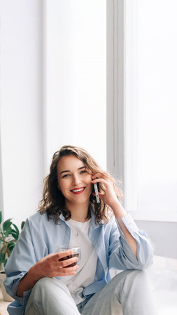 Happy woman chatting on phone with sister or friend over tea on kitchen windowsill. Pleasant conversation with loved ones. Joyful young woman having fun talking on phone while drinking tea at homeの写真素材