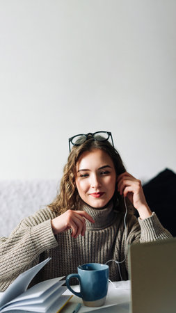 Stylish woman enjoying online education with a cup of coffee at the kitchen table, wearing headphones and writing notes while watching a webinar on her laptopの写真素材